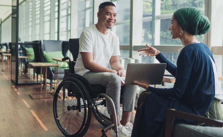 man in a wheelchair talking to a woman with a laptop